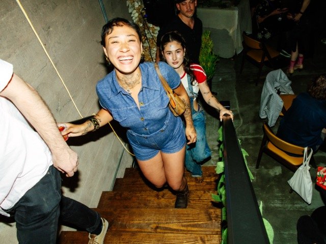 A woman with tattoos on her arms smiling as she pulls herself up a wooden staircase at a crowded indoor gathering or party.