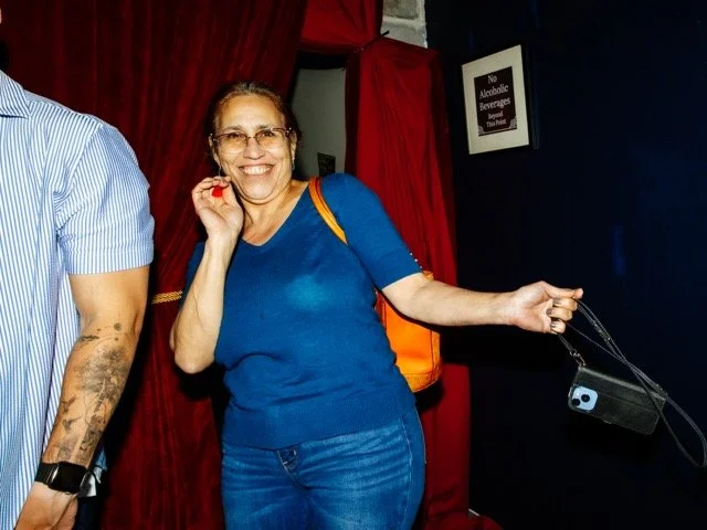 Smiling woman in blue shirt and jeans holding a camera, standing next to a person with a tattooed arm, in a dimly lit venue with red curtains and a no alcohol sign on the wall.