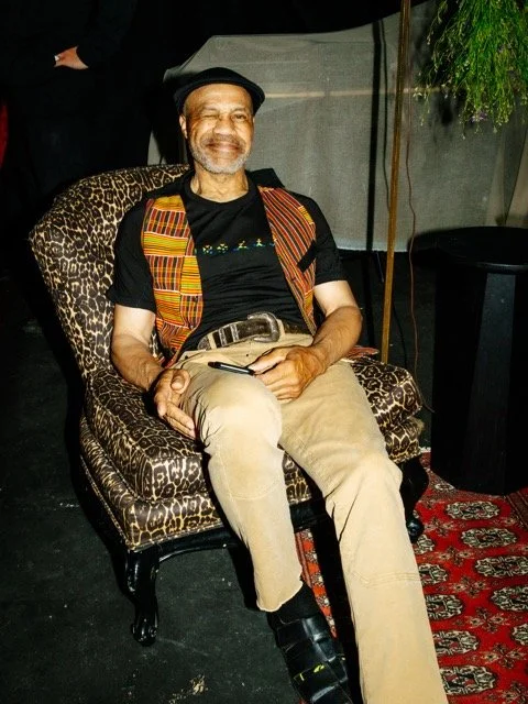 A smiling man with a beard and glasses sitting on a leopard-print chair, wearing a black cap, a black t-shirt, a colorful vest, beige pants, and black shoes, in a room with a red patterned rug and a plant.