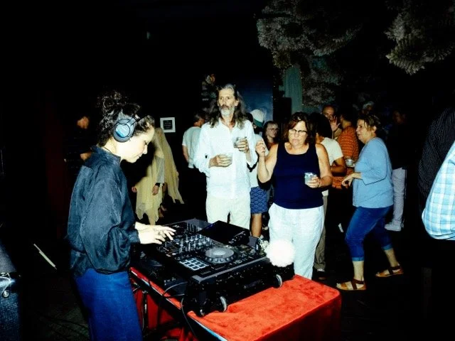 A DJ wearing headphones plays music at a party, with people dancing and socializing in the background.