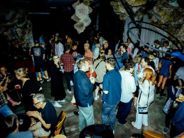 Crowd of people at a social gathering or party, some standing and chatting, others sitting at tables, in an indoor setting with a spiral staircase and decorative hanging plants.