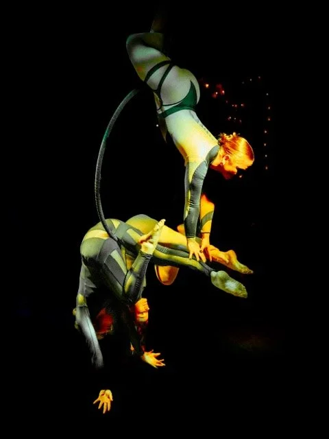 Two performers in black and gray costumes posing in an acrobatic act, one balancing upside down while the other supports them.