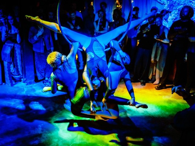 Group of performers doing acrobatic dance with colorful lighting and an audience watching.