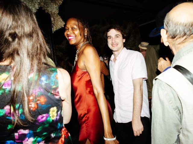Group of people at a party, smiling and socializing, in a dimly lit indoor setting.