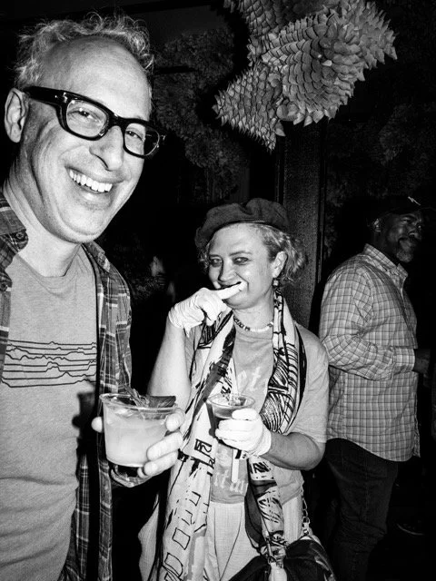 A group of people at a social gathering; a man with glasses smiling and holding a drink in the foreground, a woman with a scarf and gloves smiling and holding a drink, and a man in the background.