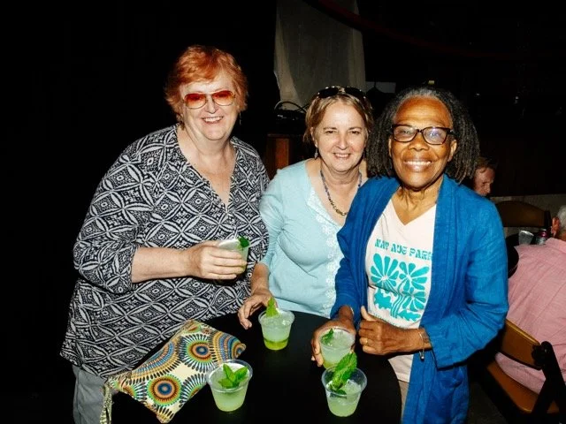 Three women smiling and holding drinks with lime garnishes at a social gathering or party.