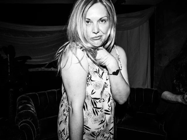 A woman with shoulder-length blonde hair wearing a floral sleeveless top, looking at the camera with a serious expression, sitting on a dark sofa in a room with draped fabric in the background.