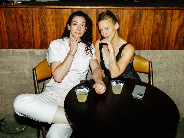 Two women sitting at a small round table with drinks, posing for the camera in a casual setting.