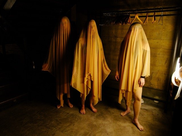 Four people covered with yellow fabric standing in a dark room, with a concrete wall and hangers in the background.