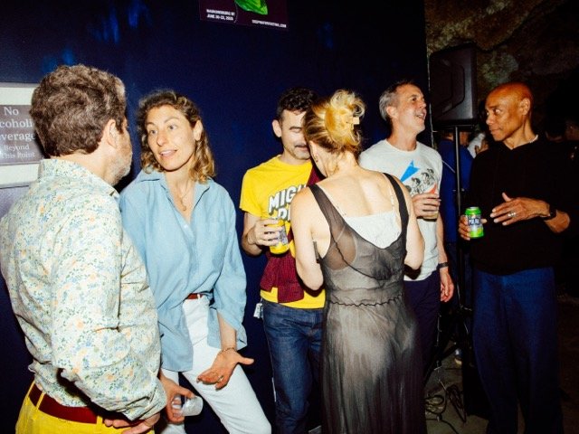 Group of six people talking and laughing at a lively social event, some holding drinks, in a dimly lit room with a blue background.