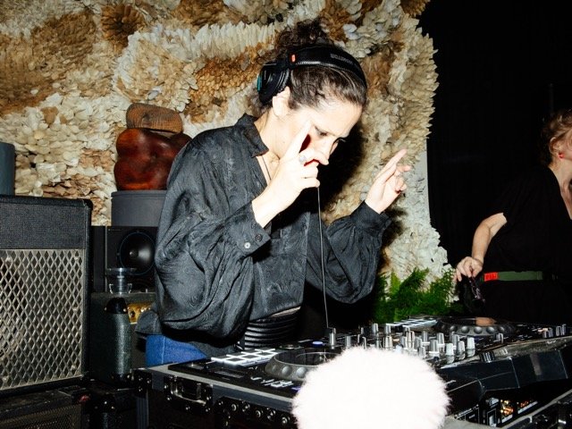A female DJ wearing headphones, with one hand near her face, operating DJ equipment in a dimly lit room with a textured wall and another person in the background.