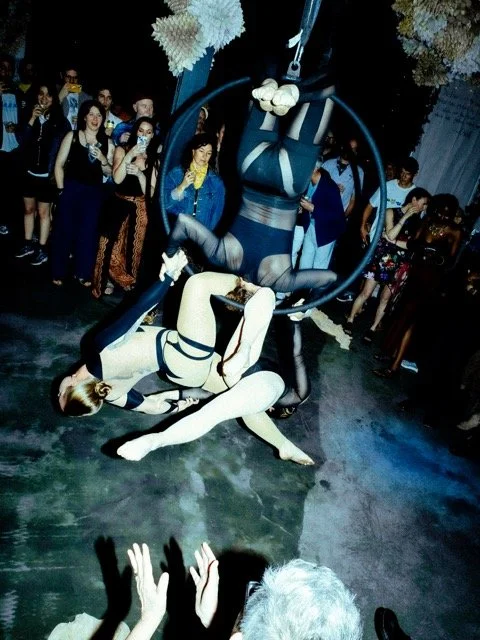 Two aerial performers in black and beige bodysuits perform acrobatics on a suspended hoop at a crowded event, audience watching and clapping.