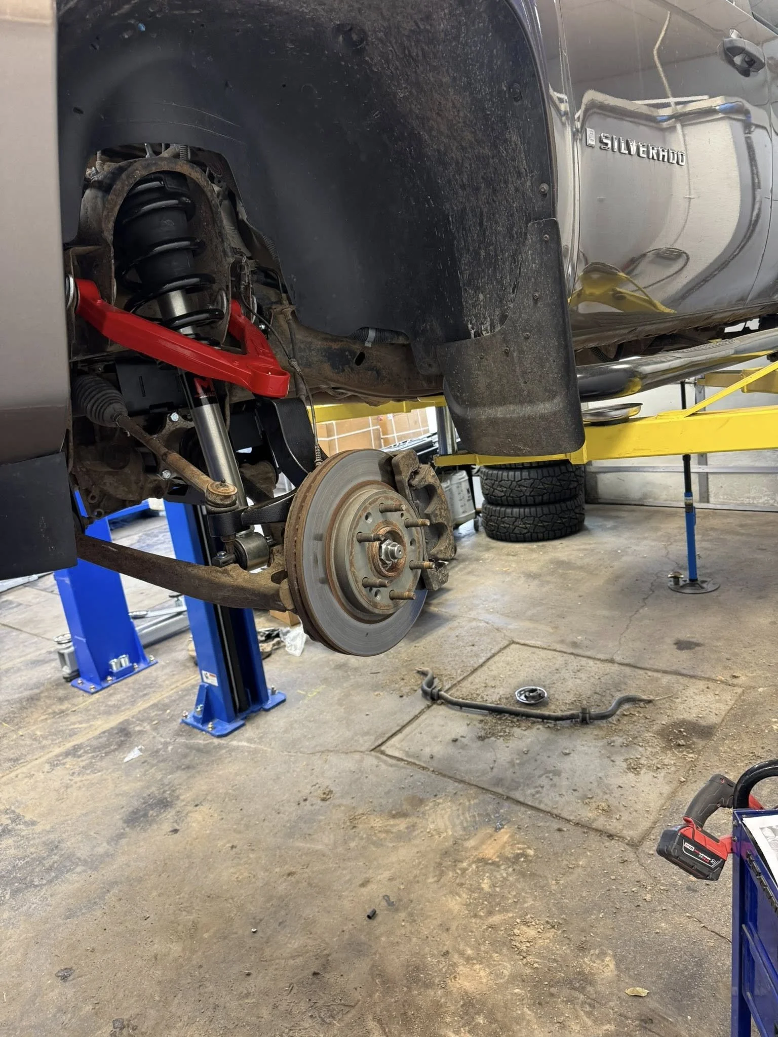 A silver Chevrolet Silverado truck raised on a blue hydraulic lift with the front wheel removed, exposing a rough country control arm on suspension lift. The truck is in an auto repair shop with tires stacked in the background and tools on the floor.