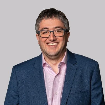 A smiling man with glasses, wearing a dark blue blazer and light pink shirt, against a light gray background.