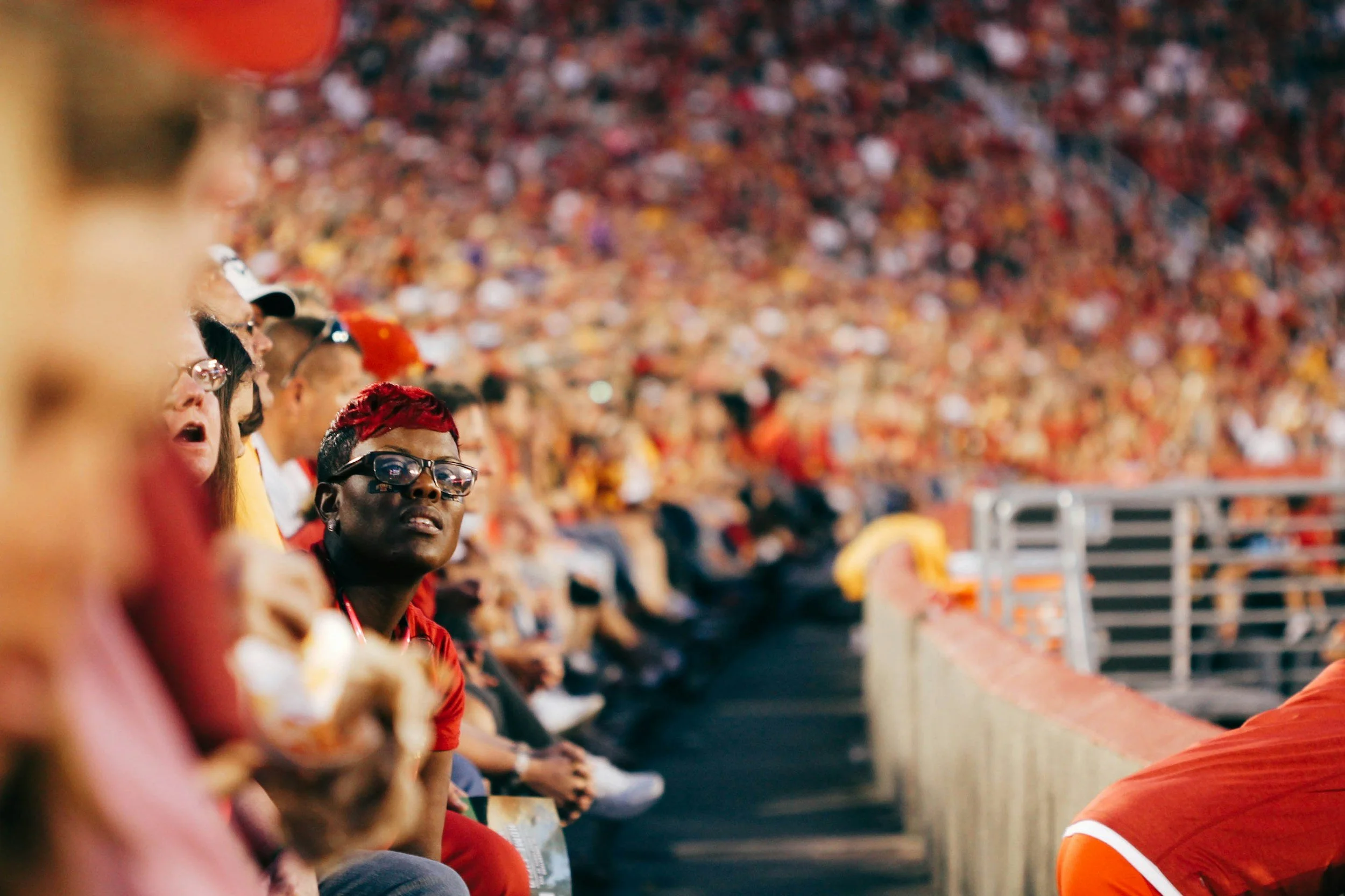 Crowd of people sitting close together in stadium seating, wearing mostly red, black, and white clothing, likely at a sports event.