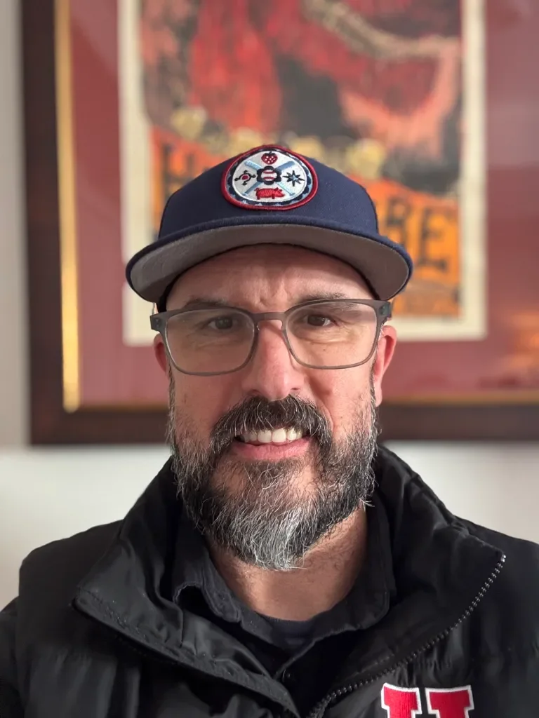 A man with glasses and a beard wearing a blue baseball cap with a colorful patch and a black jacket. Behind him is a framed abstract art piece with red, black, and orange colors.