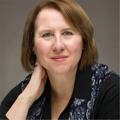 A woman with a blue collared shirt and short brown hair smiling at the camera with her hand by her face.