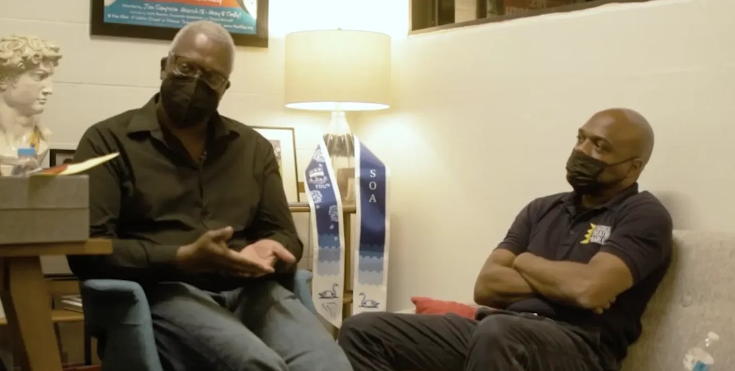 Two men sitting and talking in a room with framed pictures on the wall, a lamp, and a sculpture bust. Both men are wearing black face masks.