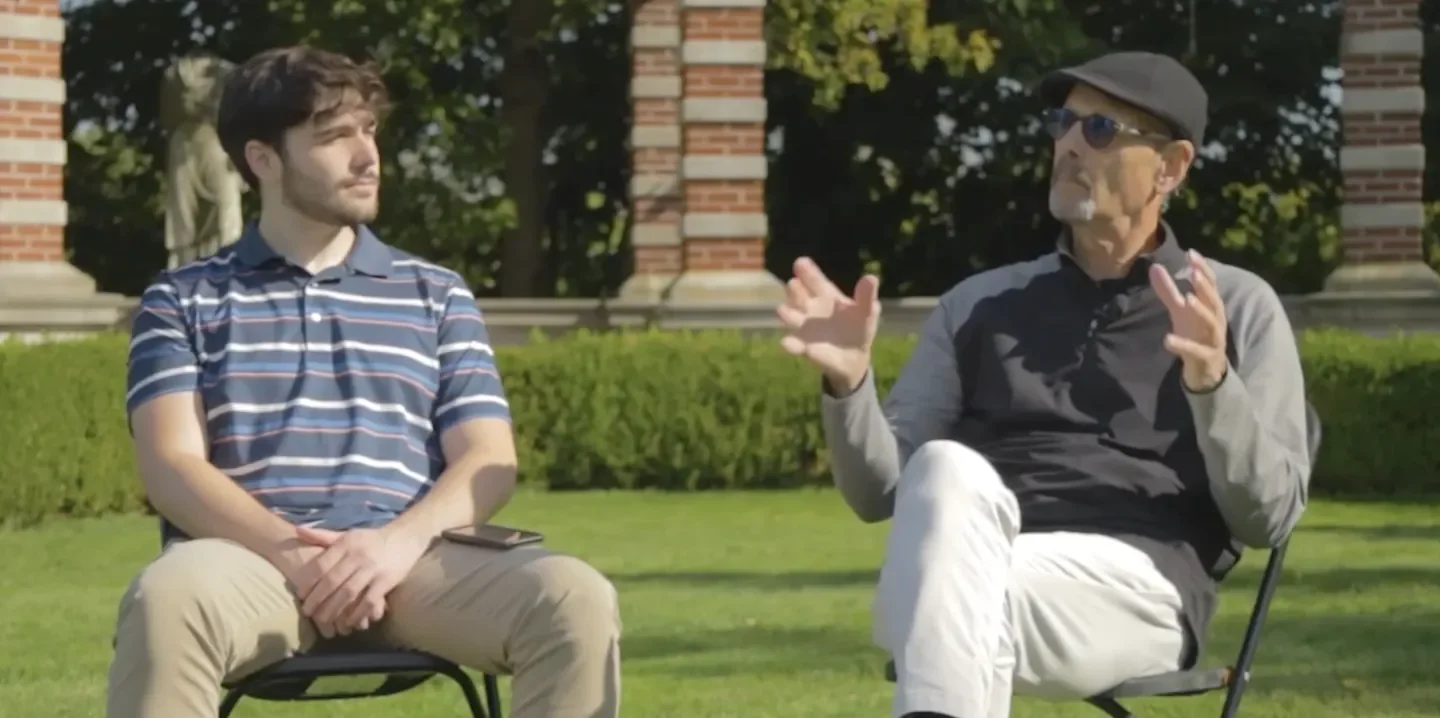 Two men sitting outside on chairs engaging in a conversation on a sunny day, with a brick monument and trees in the background.