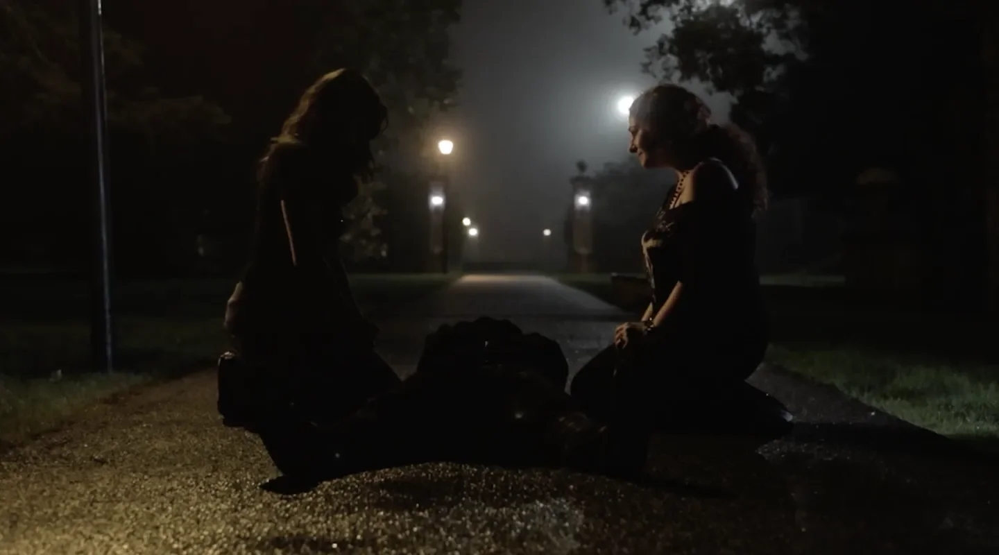 Two women kneeling on a dark, foggy street at night, illuminated by streetlights, with their heads bowed and hands clasped, appearing to pray or have a serious conversation.