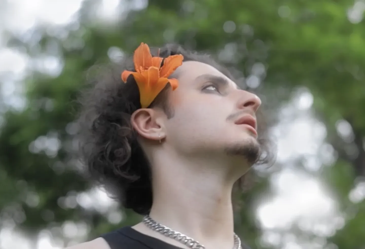 Person with long curly hair, wearing a silver chain necklace, has an orange flower tucked behind their ear, standing outdoors with a blurred green background.