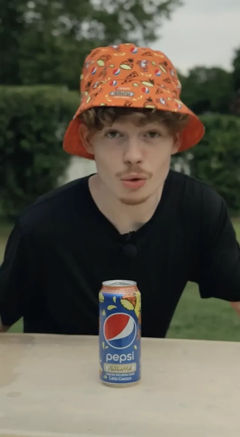 A young man with curly hair and a mustache wearing an orange bucket hat with colorful snack and soda illustrations, leaning over a table with a Pepsi soda can in front of him outdoors with greenery in the background.