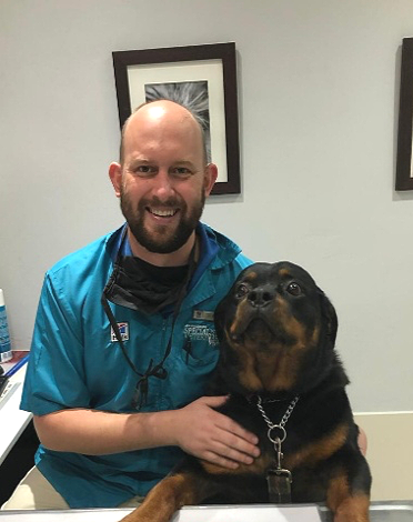 A smiling man with a beard wearing blue scrubs is sitting at a desk with a Rottweiler dog. The man has his hand on the dog, and the background shows framed artwork on a gray wall.