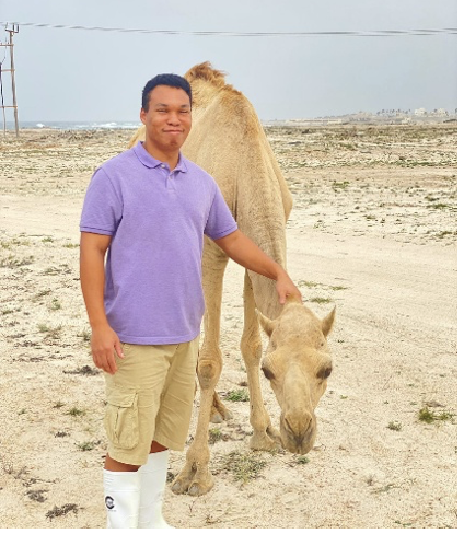 A young man in a purple polo shirt and beige shorts standing next to a light-colored camel in a barren, open landscape.