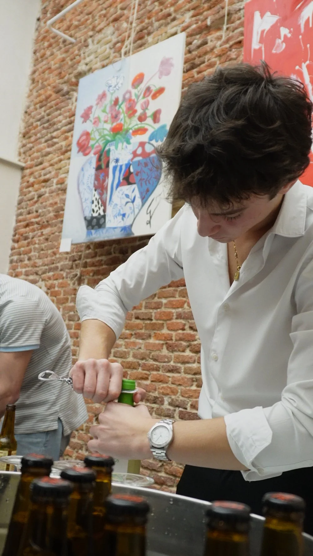 Hombre abriendo una botella en una mesa con varias botellas pequeñas, en un fondo de pared de ladrillos y una pintura colorida de un jarrón con flores.