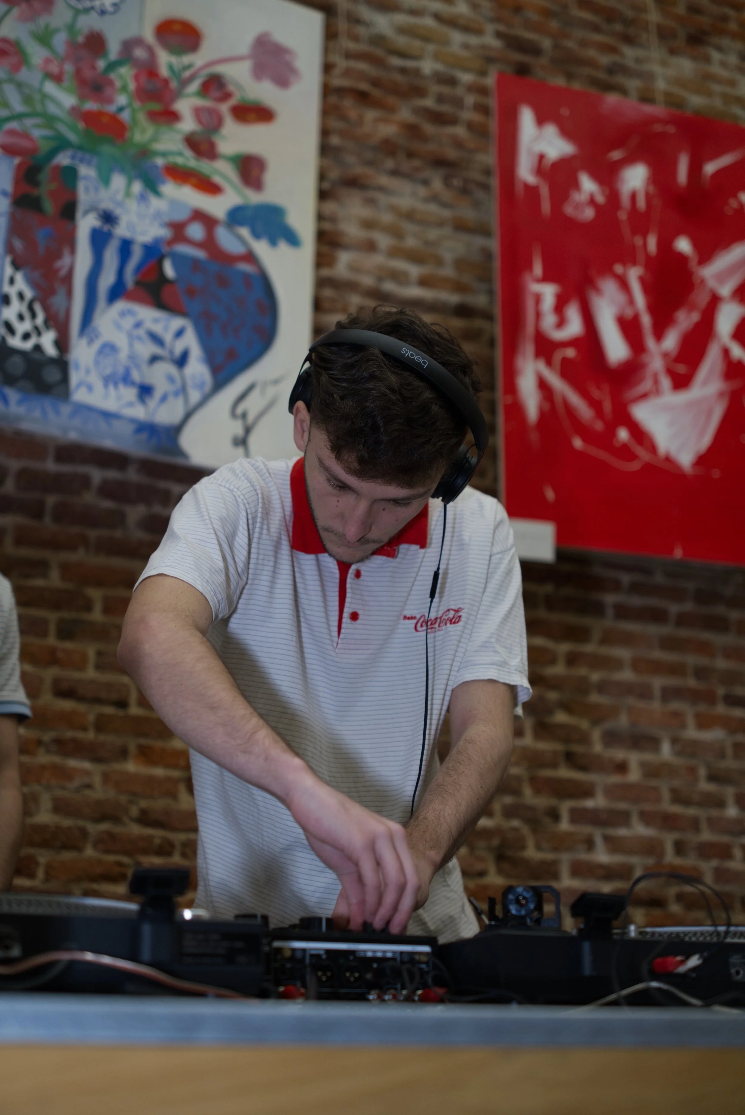 Joven DJ mezclando música en un equipo de sonido en un evento, con paredes de ladrillo y arte moderno en el fondo.
