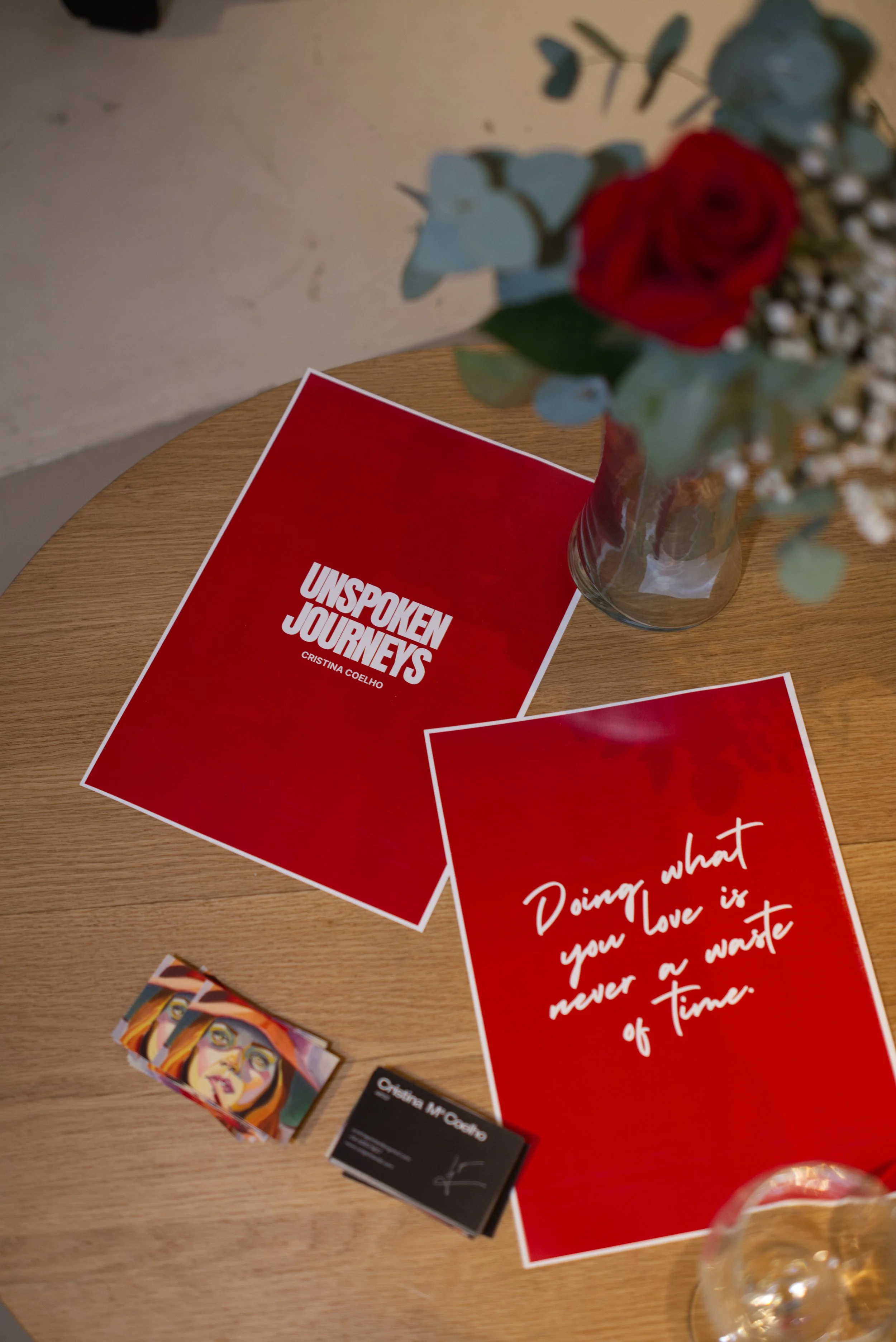 Una mesa de madera con un libro titulado 'Unspoken Journeys' de Cristina Coelho, una tarjeta con la frase 'Doing what you love is never a waste of Time', un pequeño conjunto de tarjetas de visita, un florero con flores rojas y verdes, y una copa de v