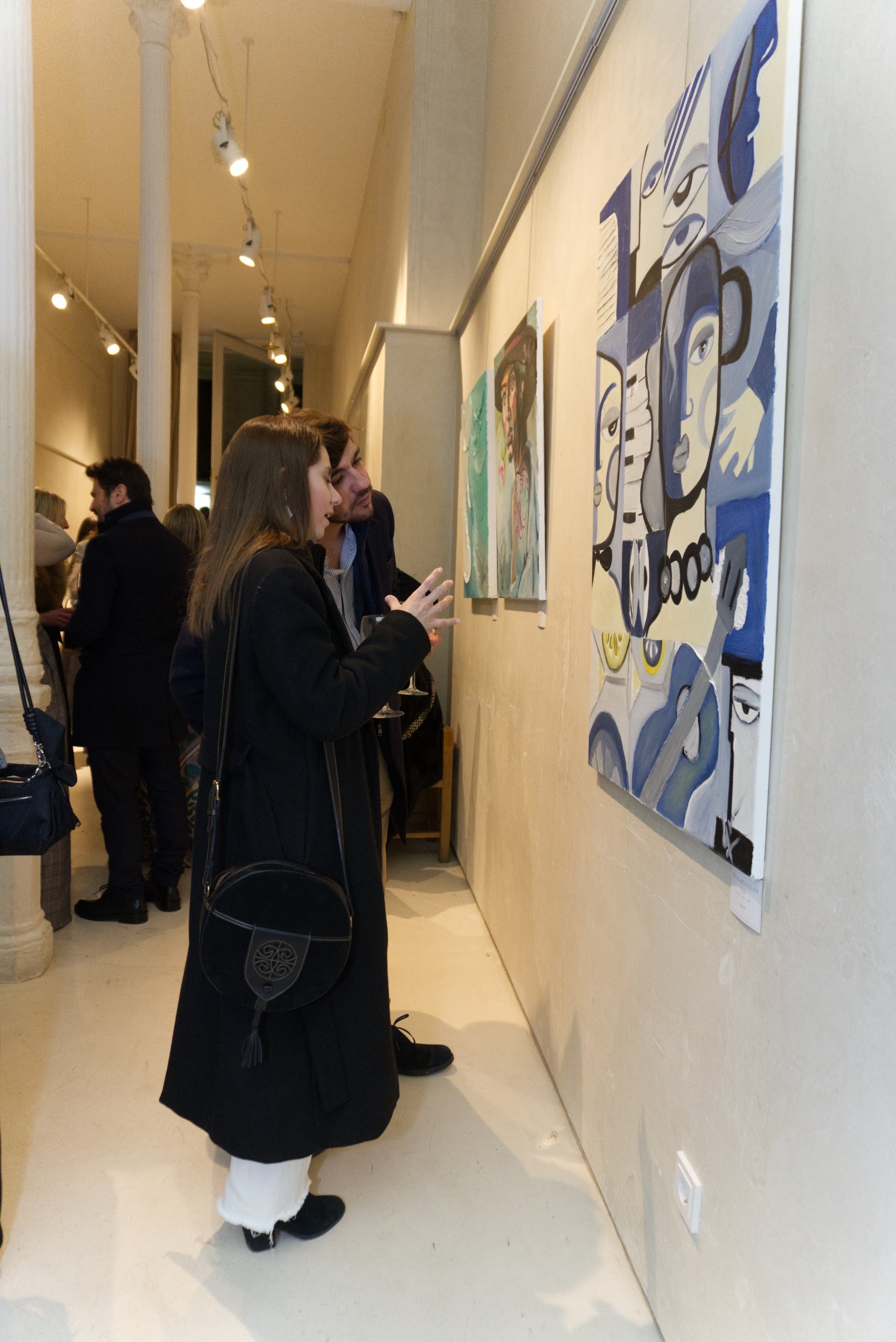 Personas observando cuadros en una galería de arte, iluminada con focos en el techo.