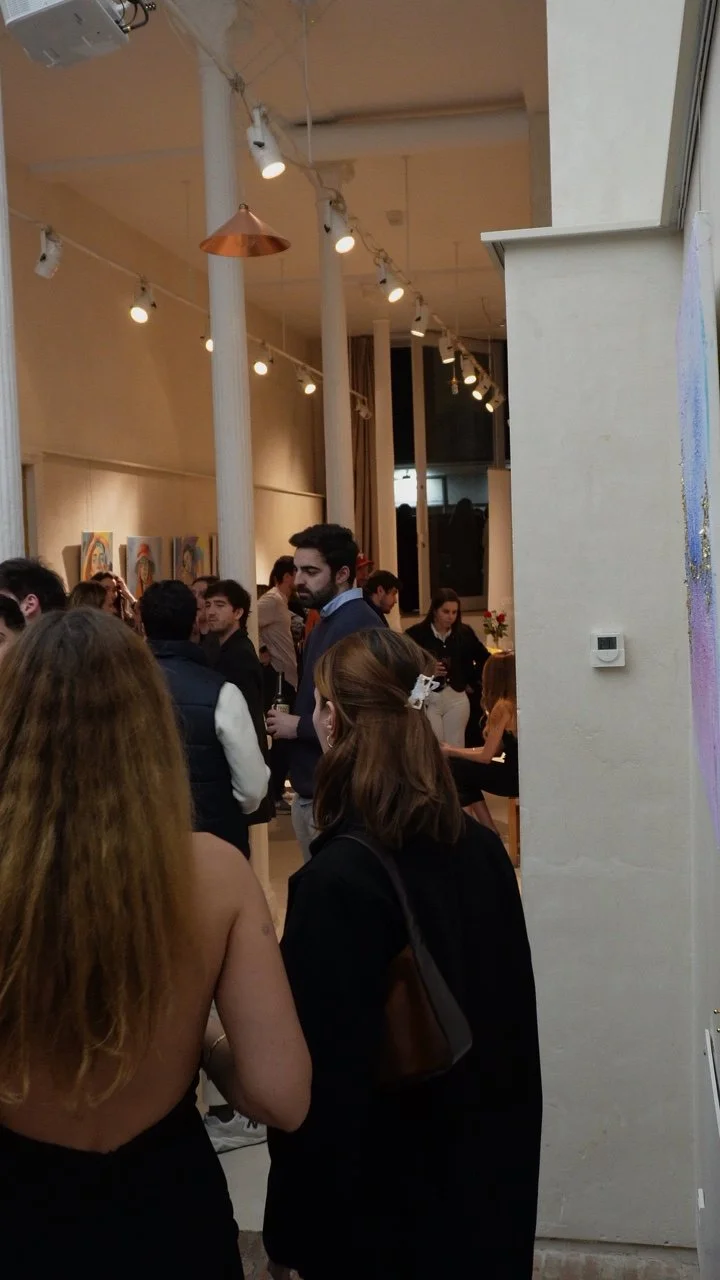 Una reunión en un espacio interior con varias personas conversando, decoraciones y pinturas en las paredes, iluminación con luces colgantes y columnas blancas.