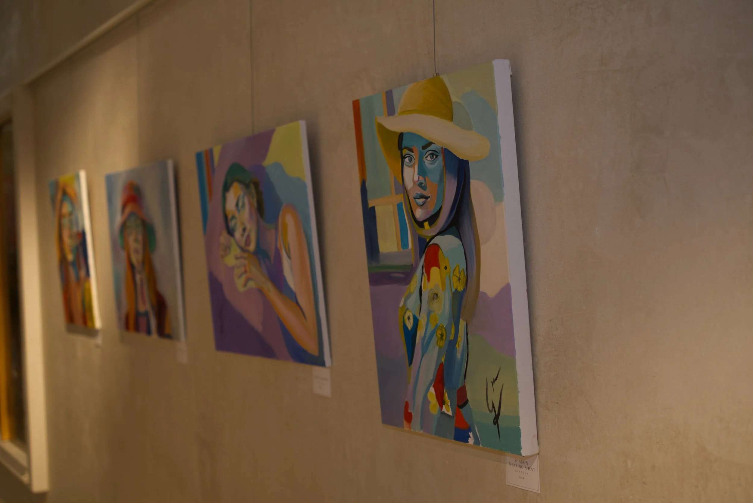 Cuadros de arte en una galería, con retratos en estilo moderno. La imagen en primer plano muestra a una mujer con sombrero amarillo, cabello largo y ropas coloridas.