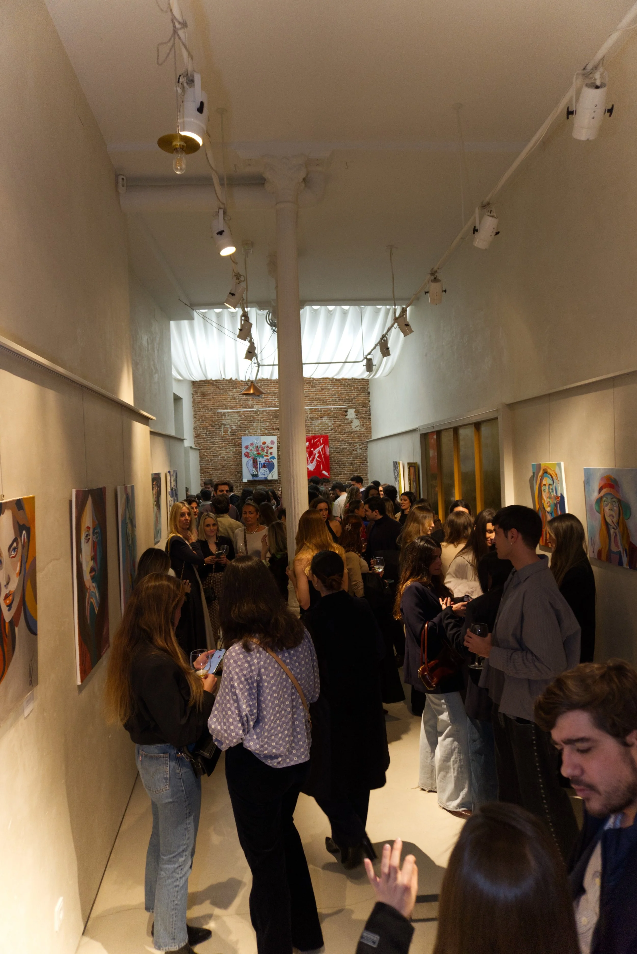 Vista de una galería de arte con muchas personas observando pinturas en las paredes, con iluminación dirigida y una pared de ladrillo en el fondo.