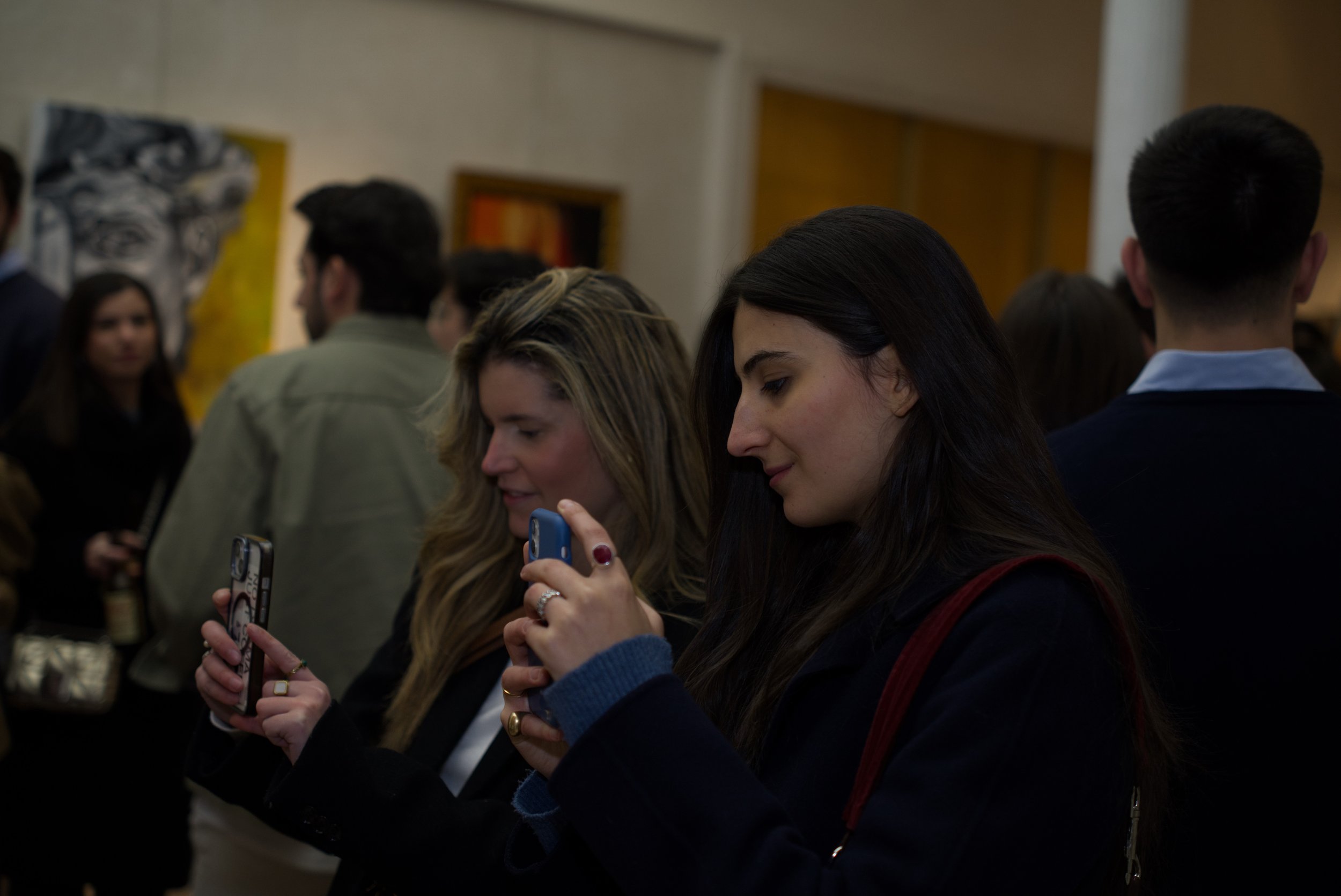 Personas en un evento cultural toman fotos con teléfonos inteligentes en un espacio con pintura artística en la pared.