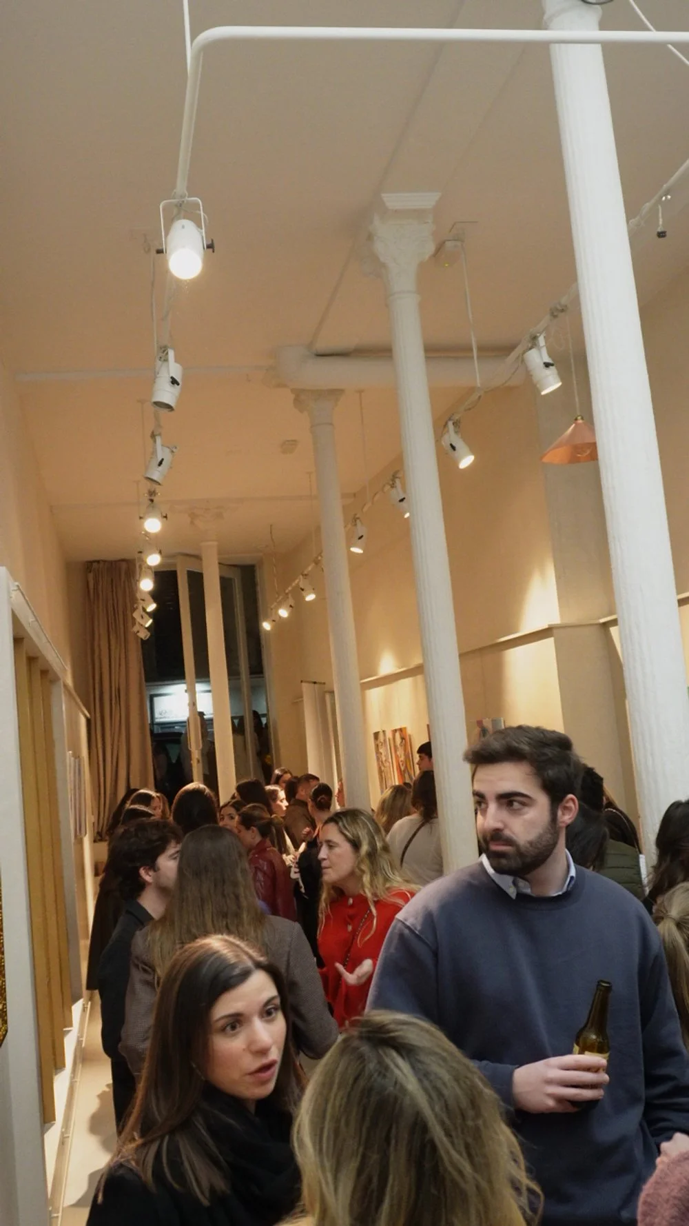 Grupo de personas en una sala de exposición de arte, con columnas blancas y luces de techo apuntando hacia las obras en las paredes.