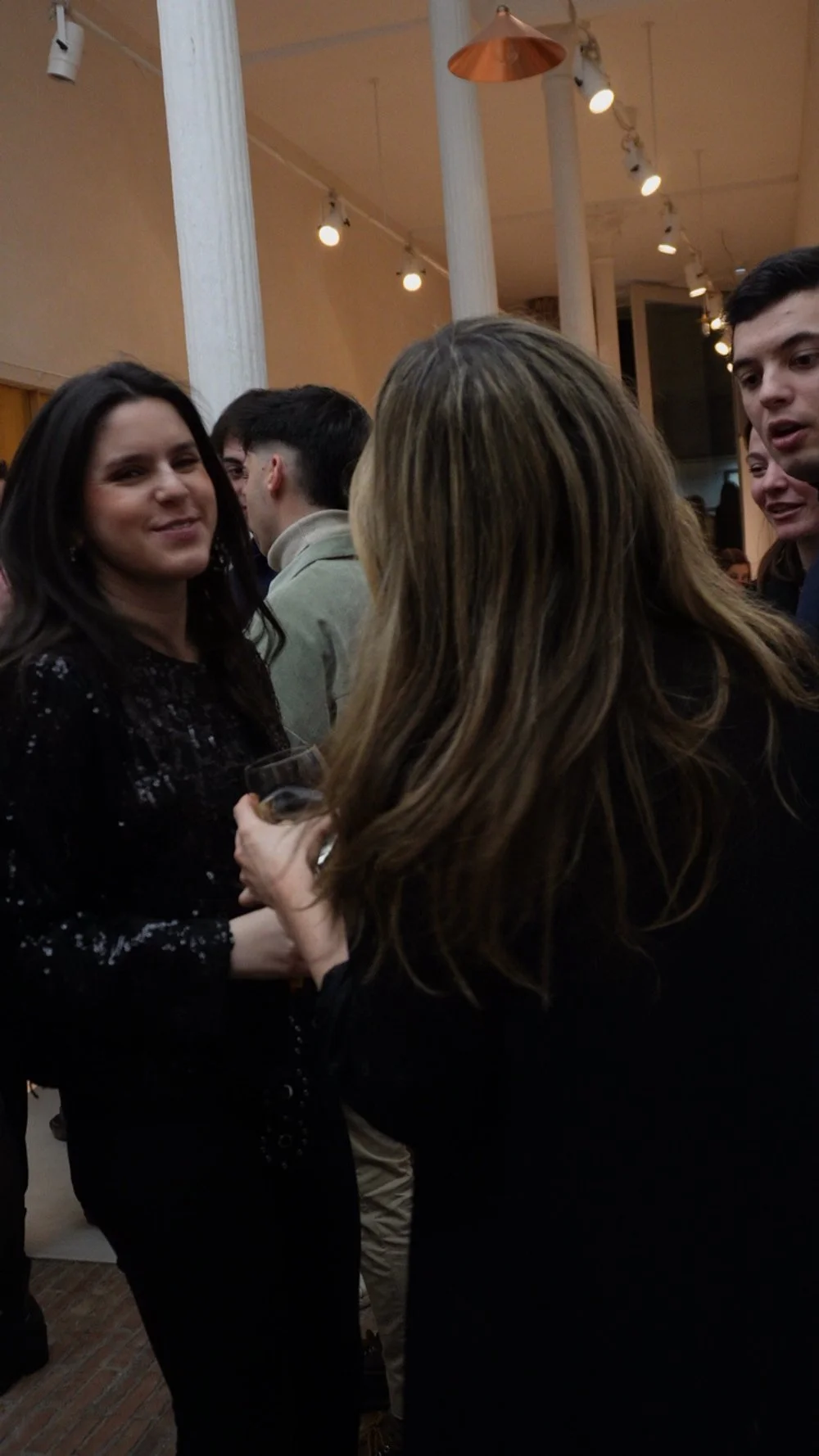 Grupo de personas en una reunión social en un lugar decorado con luces y columnas de madera, conversando y disfrutando del momento.