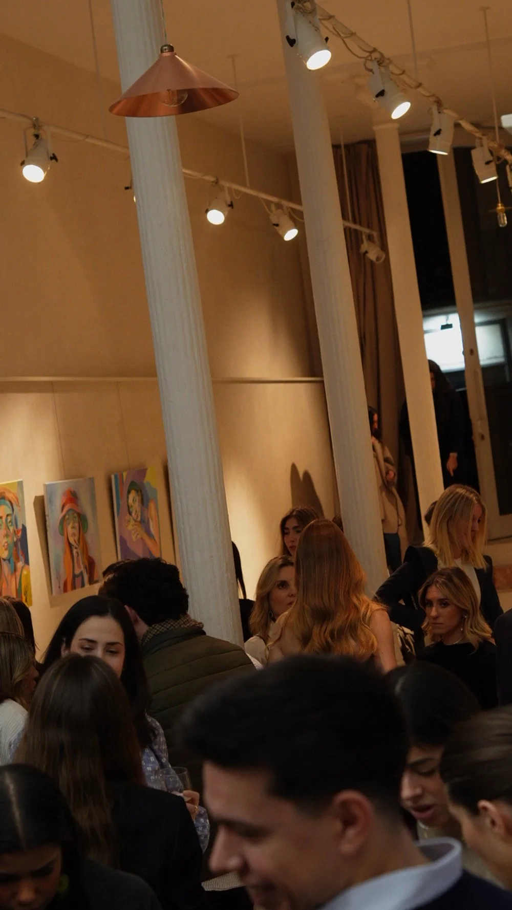 Grupo de personas en una galería de arte moderna, con paredes decoradas con retratos coloridos, en un ambiente bien iluminado con iluminación fija y columnas blancas.