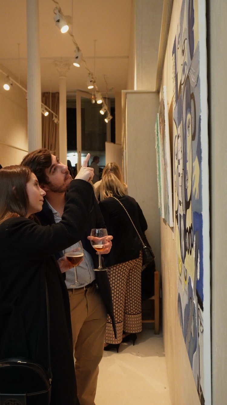 Personas observando y comentando obras de arte en una galería, con copas en las manos, en un ambiente de exposición.
