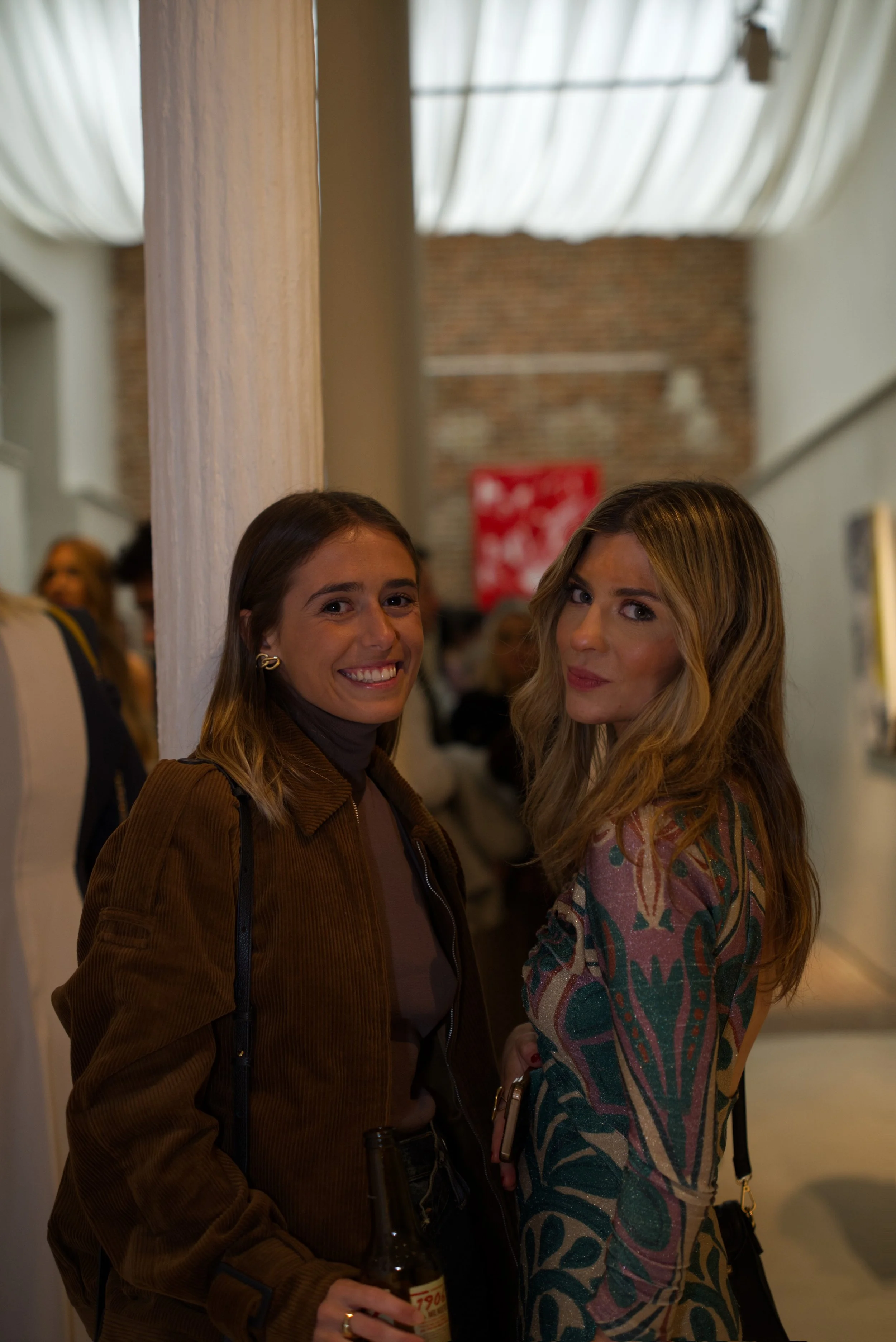 Dos mujeres adultas posando dentro de un espacio interior con paredes de ladrillo, una de ellas sostiene una botella y ambas miran hacia la cámara, en un ambiente que parece ser una galería o evento social.