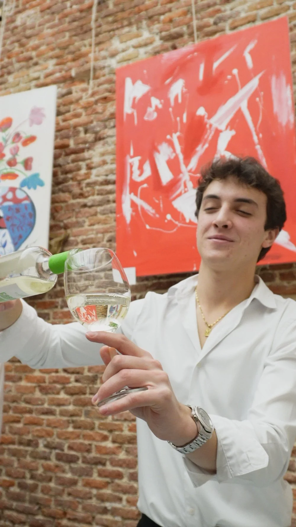 Joven sirviendo vino en una copa en un espacio con paredes de ladrillo y obras de arte coloridas.