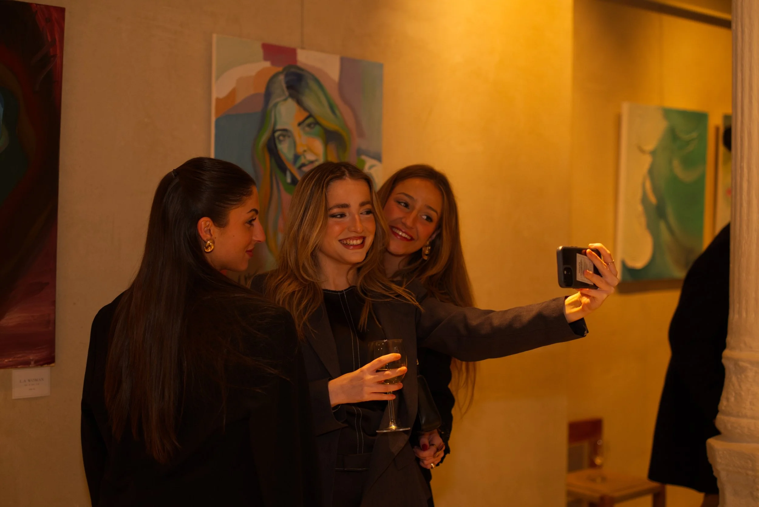 Tres mujeres jóvenes en una galería de arte, una de ellas toma una selfie mientras sostienen copas de vino, con pinturas en el fondo.