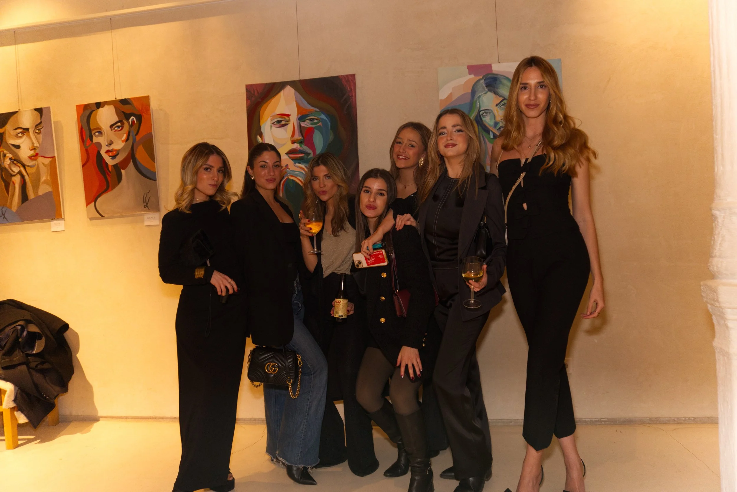 Grupo de siete mujeres en una galería de arte posando en frente de cuadros abstractos de retratos de mujeres.