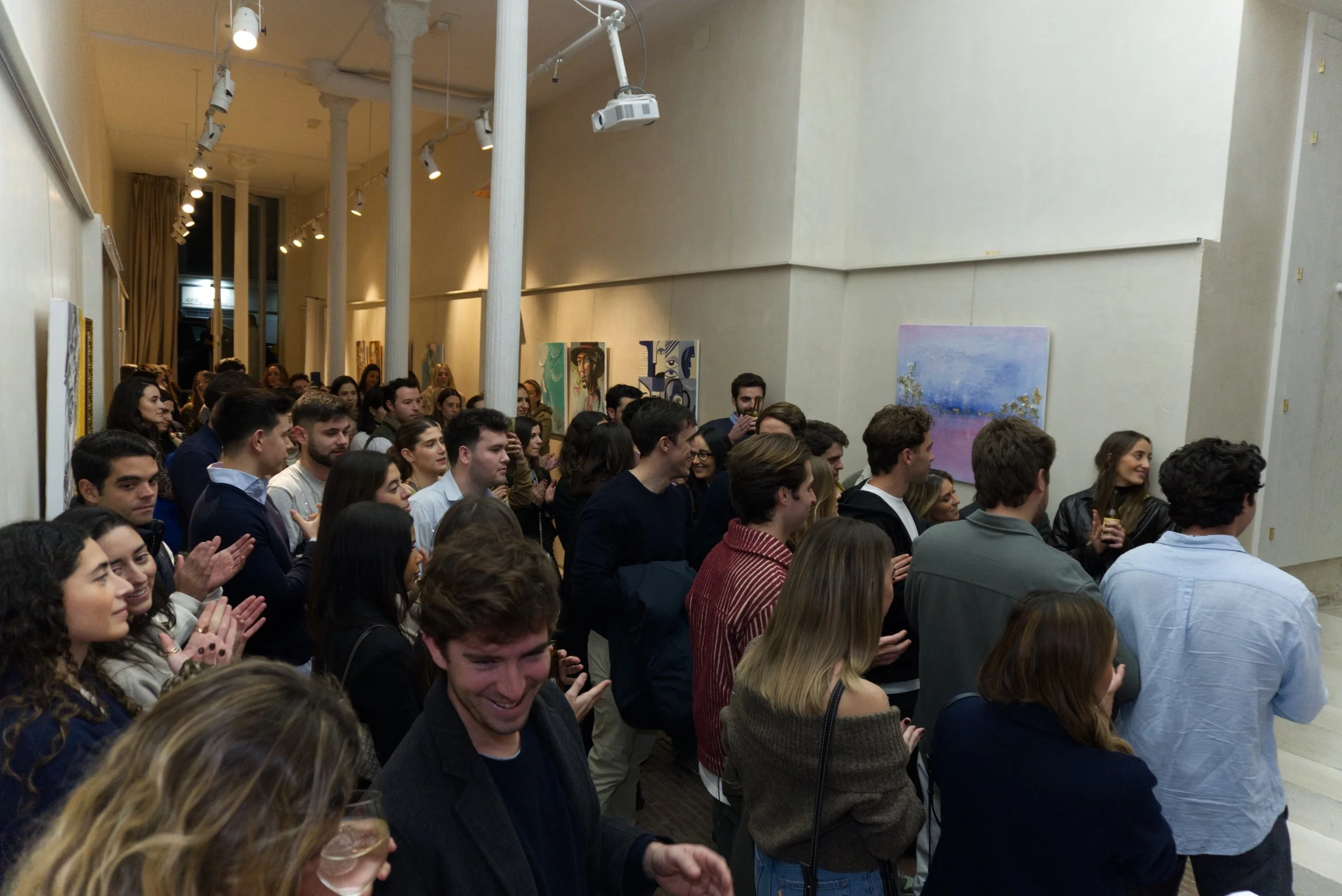 Grupo de personas en una sala de arte durante una inauguración o evento, observando pinturas en las paredes, algunos aplaudiendo y otros tomando fotos.