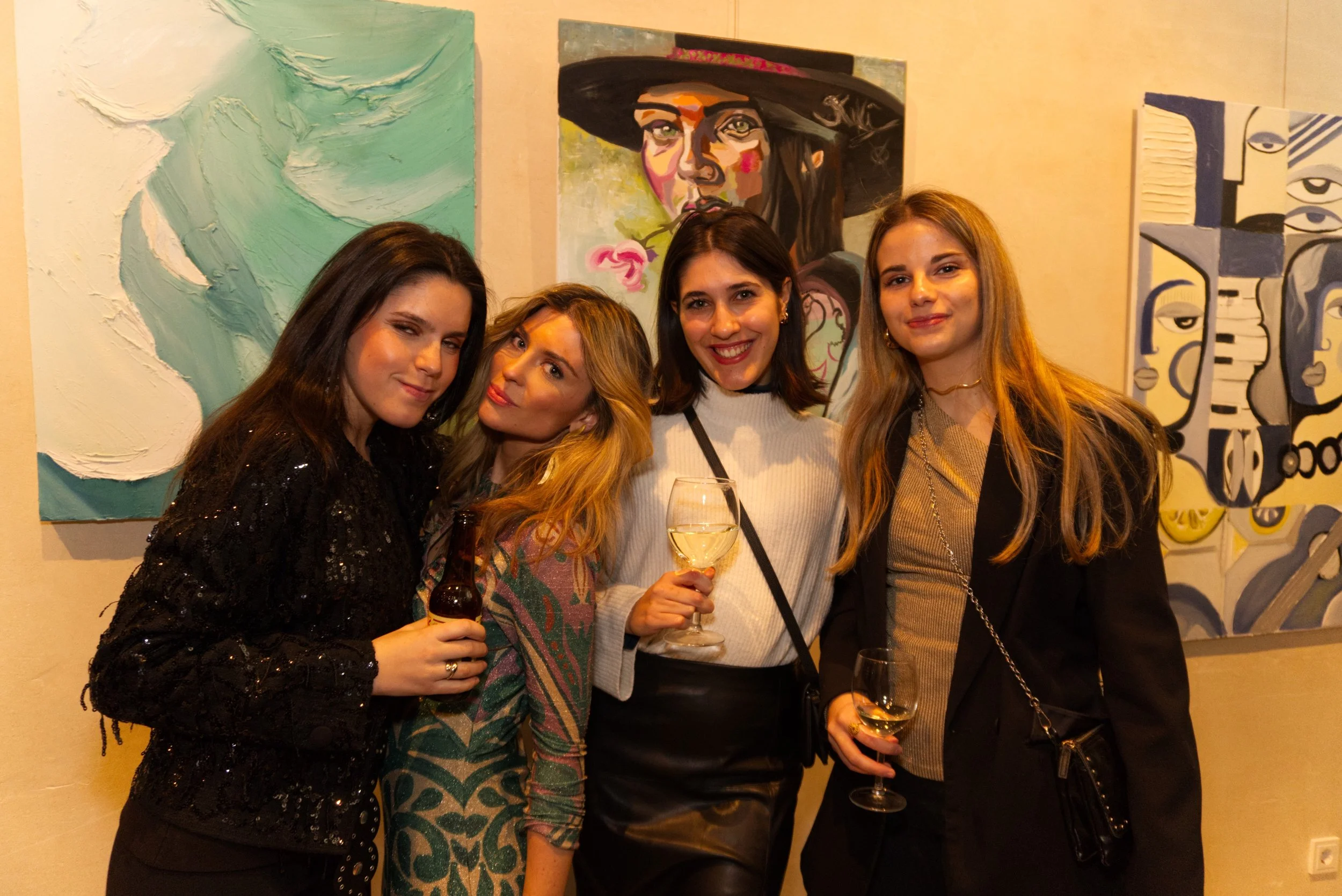 Cuatro mujeres en una galería de arte posando con pinturas abstractas en el fondo, algunas con bebidas en la mano, sonrientes y disfrutando del evento.