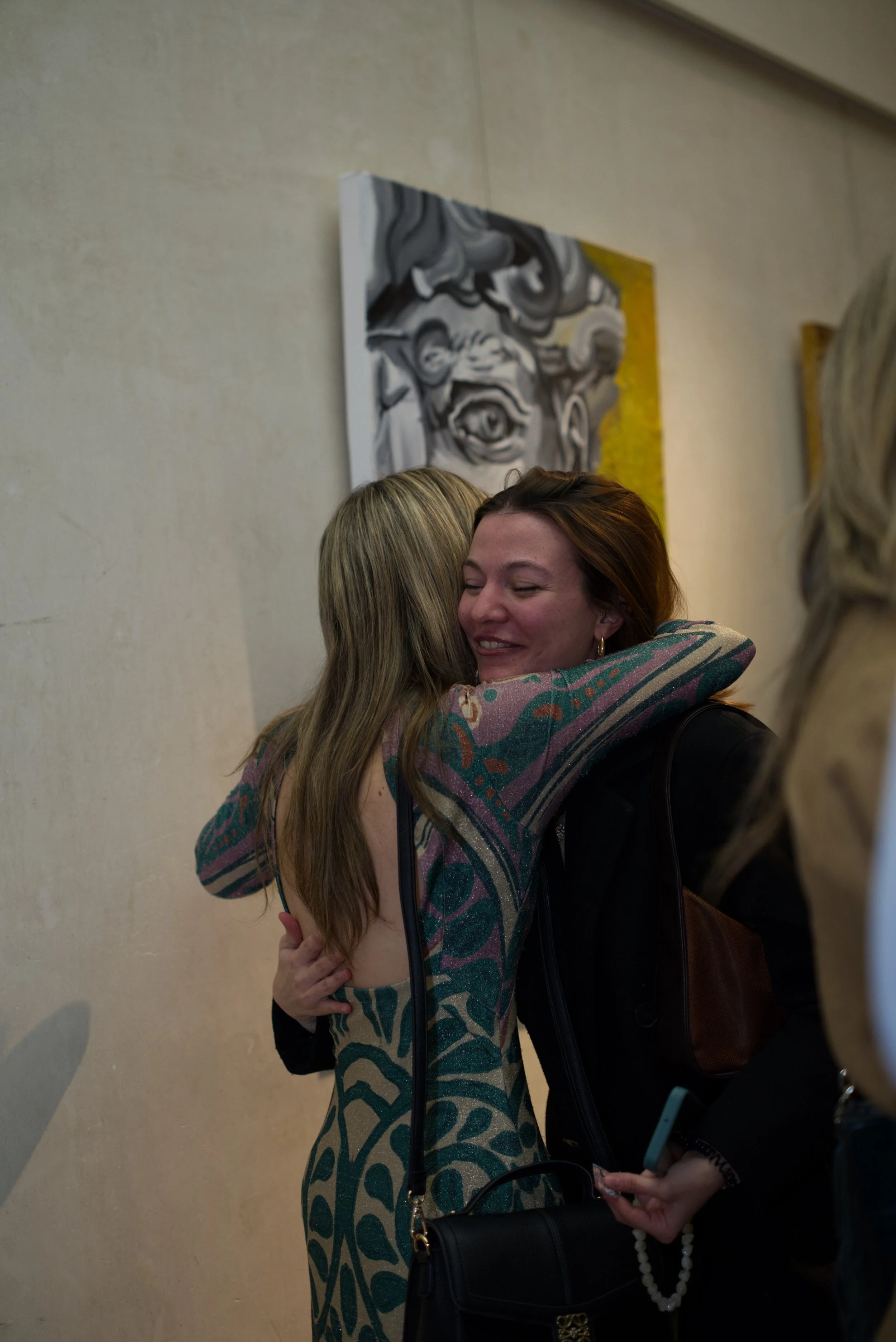 Dos mujeres abrazándose y sonriendo en un evento, con una pintura abstracta en gris y amarillo en la pared de fondo.