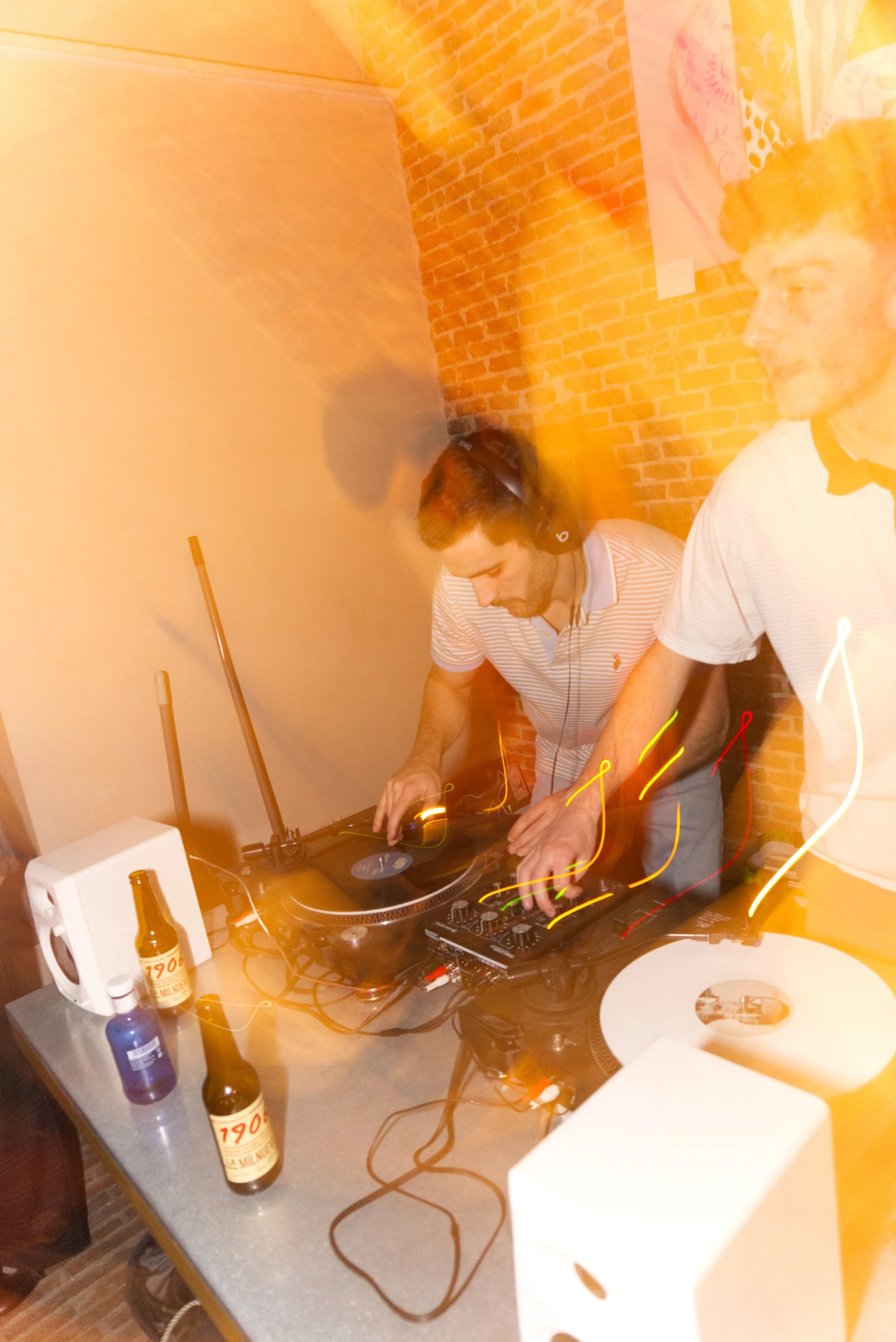 Dos personas mezclando música en un equipo de DJ en una habitación con pared de ladrillos expuestos, botellas en la mesa, y luces de colores movidas.