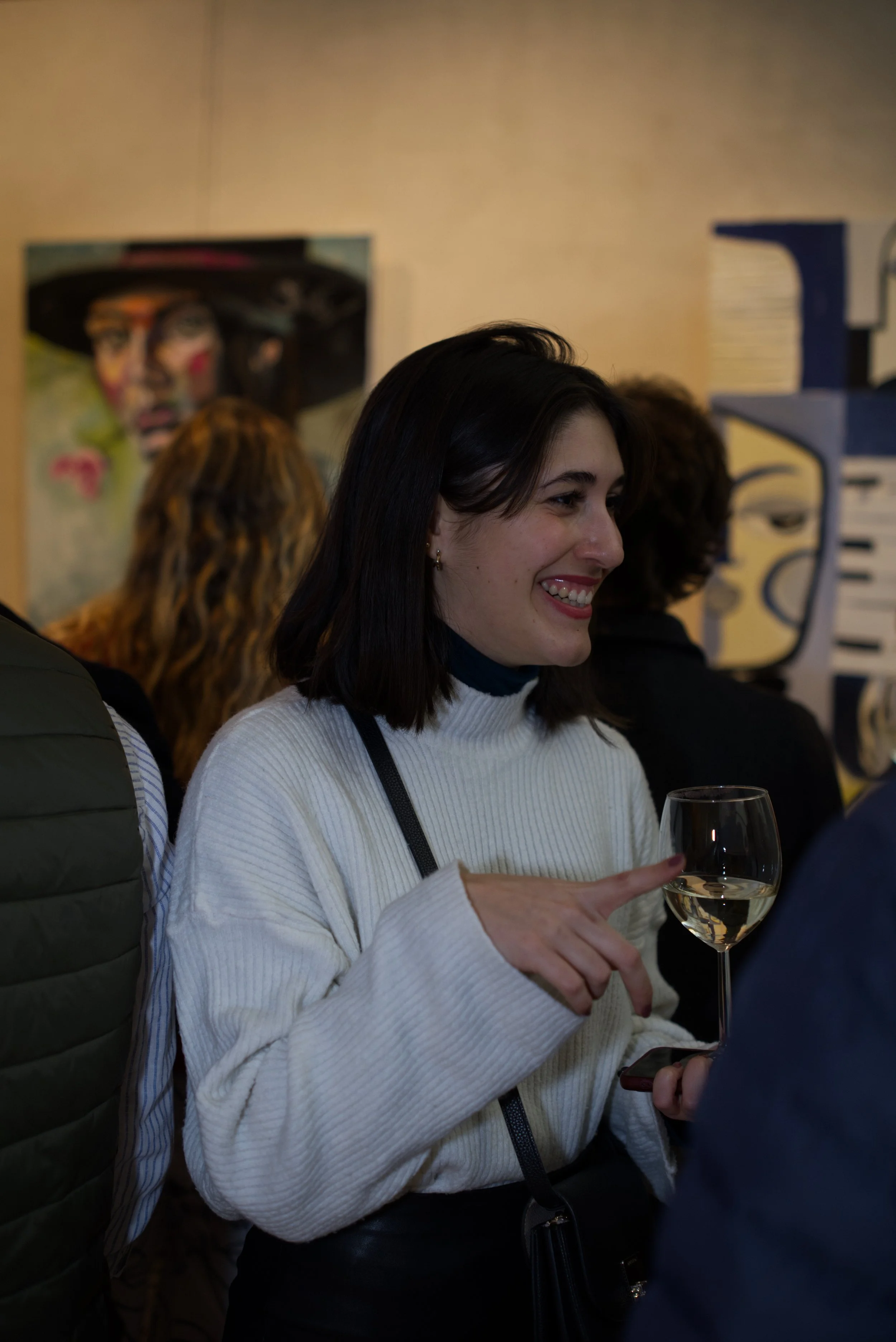 Mujer sonriendo y sosteniendo un vaso de vino en un evento social, con fondo de arte y otras personas.