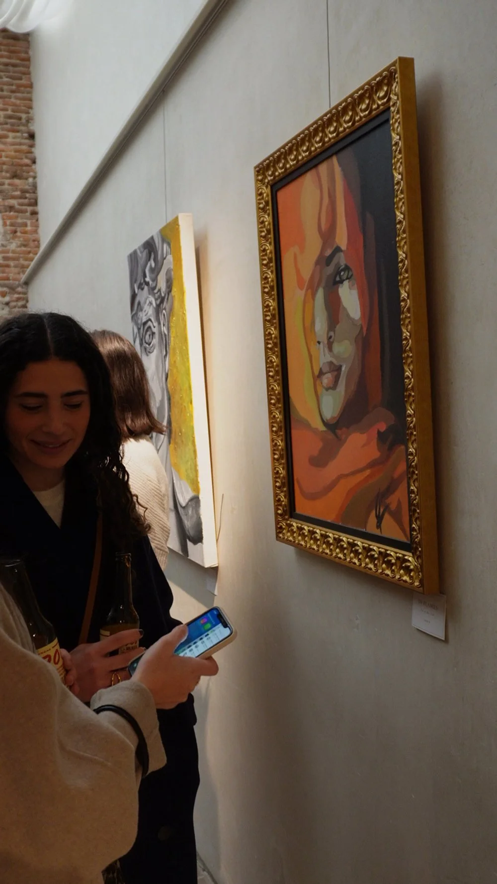 Personas observando pinturas en una galería de arte, una de ellas sostiene una botella y un celular